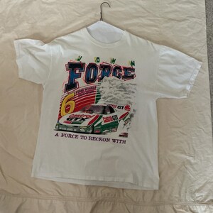 May include: A white short-sleeve t-shirt featuring a graphic of a John Force race car with Castrol GTX branding. The design includes the text FORCE, 16 TIME WORLD CHAMPION, and A FORCE TO RECKON WITH, along with a large yellow number 6.