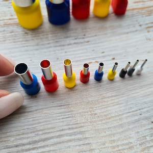 Peut inclure: Assortiment de connecteurs de fil de différentes tailles, de couleurs vives, avec un doigt tenant le plus grand connecteur. Les connecteurs sont en plastique et ont un anneau métallique sur le dessus.