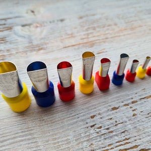 Pode incluir: Um conjunto de dez bicos de pastelaria de metal de diferentes tamanhos e formas. Os bicos estão dispostos por ordem de tamanho, com o bico maior à esquerda e o menor à direita. Os bicos são todos de cor prateada e têm um acabamento liso e polido. Os bicos estão presos a uma pega de plástico que é amarela, azul ou vermelha.