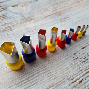 May include: A set of ten metal piping tips with different shaped openings. The tips are arranged in a row on a light wood surface. The tips are color-coded with red, yellow, and blue.
