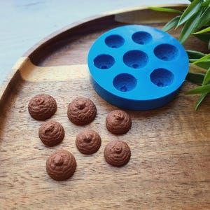 May include: A blue silicone mold with eight round cavities, used for making small, round chocolate candies. Eight brown chocolate candies are shown on a wooden surface next to the mold.
