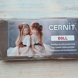 May include: A block of brown modeling clay with a porcelain finish. The packaging features two young girls wearing white dresses and red flower crowns. The text on the packaging reads "CERNIT DOLL Modelling clay with porcelain finish Pate à modeler finition porcelaine Modelliermasse mit Porzellanschimmer".