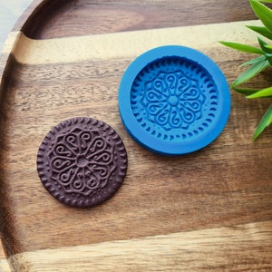 Peut inclure: Un biscuit rond brun foncé à côté d'un moule en silicone bleu avec un motif floral détaillé. Le biscuit et le moule sont sur un plateau en bois, avec un feuillage vert en arrière-plan. Le biscuit a un motif détaillé.