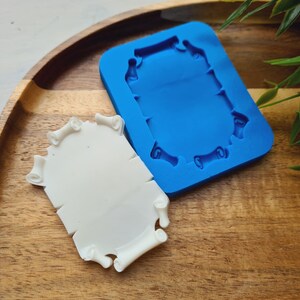 Op de afbeelding: Een wit, rolvormig object naast een blauwe siliconen mal op een houten dienblad. De mal heeft een bijpassende rolvorm. Groene bladeren zijn op de achtergrond. De objecten zijn waarschijnlijk voor knutselen of het maken van decoratieve items.