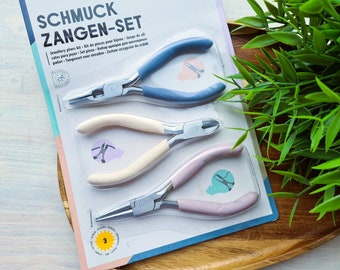 Puede incluir: Un kit de alicates para joyería con tres alicates diferentes. Los alicates tienen cabezas de metal plateado y mangos de colores en azul, crema y rosa. El embalaje incluye texto en varios idiomas, incluyendo "SCHMUCK ZANGEN-SET".