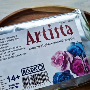 May include: A package of white air-drying clay, labeled "Artista Extremely Lightweight Airdrying Clay" with a 170 gram weight. The package features a 14+ age recommendation and a barcode. The clay is made by PADICO and is suitable for crafting projects.