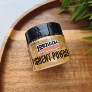 Könnte beinhalten: Ein kleines Glas Goldpigmentpulver mit schwarzem Deckel, beschriftet mit "pentart PIGMENT POWDER". Das Glas steht auf einem Holztablett, im Hintergrund grüne Blätter. Das Pigment scheint ein feines, schimmerndes Pulver zu sein.