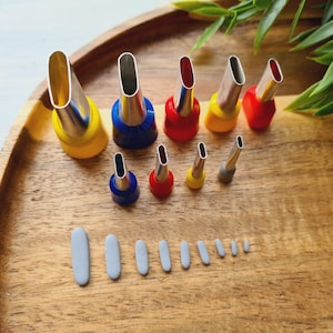 Può includere: Una collezione di nove beccucci in metallo con basi in plastica colorata gialla, blu e rossa, disposti su due file. Sotto i beccucci ci sono nove piccoli pezzi di argilla o plastica grigia di forma ovale e di dimensioni variabili.