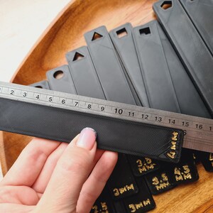 Peut inclure: Une r&egrave;gle noire avec une surface textur&eacute;e est plac&eacute;e sur un ensemble d'outils de mesure noirs. La r&egrave;gle est gradu&eacute;e en pouces et en centim&egrave;tres. Plusieurs outils de mesure noirs sont visibles, avec du texte dor&eacute; indiquant les mesures en millim&egrave;tres.