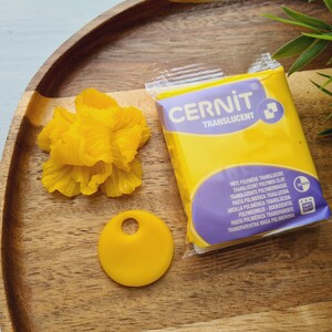 May include: A wooden tray holds a yellow Cernit translucent polymer clay package, a yellow flower, and a yellow pendant with a hole. The clay package has purple and white text. Green leaves are in the background.