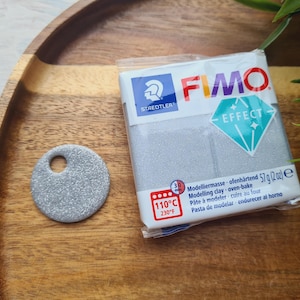 May include: A package of FIMO Effect modeling clay, 2 oz, next to a round, silver-colored pendant with a hole. The package has the Staedtler logo and a diamond-shaped graphic. The clay is oven-bake, at 230°F (110°C).