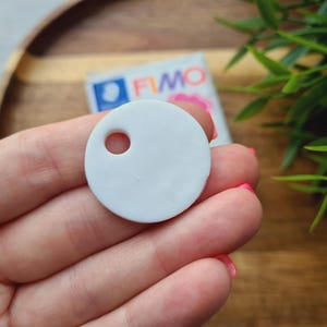 May include: A light gray, round polymer clay pendant with a hole, held in a hand. The pendant is smooth and matte. A package of FIMO polymer clay is visible in the background. The pendant is likely for jewelry making or crafting.