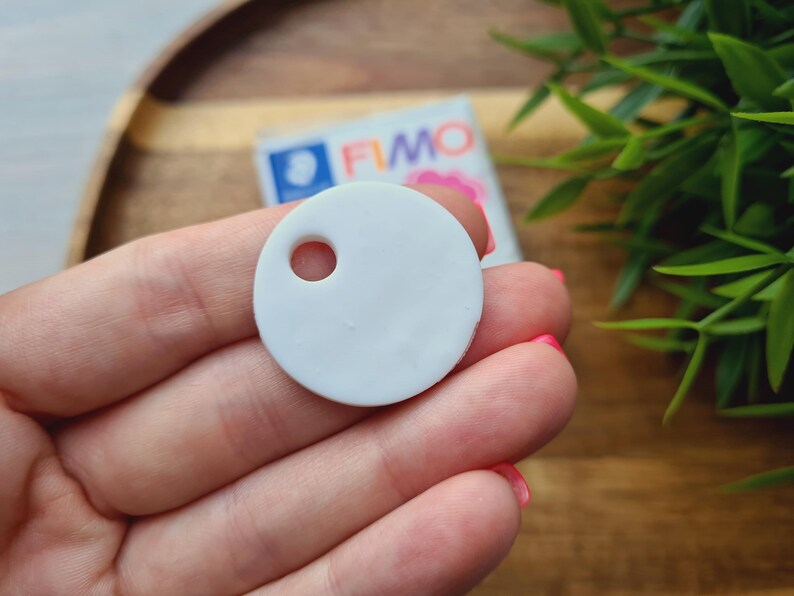 May include: A light gray, round polymer clay pendant with a hole, held in a hand. The pendant is smooth and matte. A package of FIMO polymer clay is visible in the background. The pendant is likely for jewelry making or crafting.