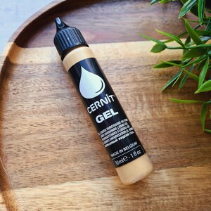 May include: A black and tan tube of CERNIT GEL, a liquid hardening gel, with a white drop logo. The tube is 1 fl oz and made in Belgium. The product is on a wooden tray.