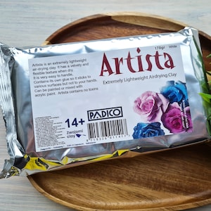 May include: A silver package of white air-drying clay labeled "Artista Extremely Lightweight Airdrying Clay" with a barcode and the text "170gr White". The package features a floral design with pink, blue, and purple roses.