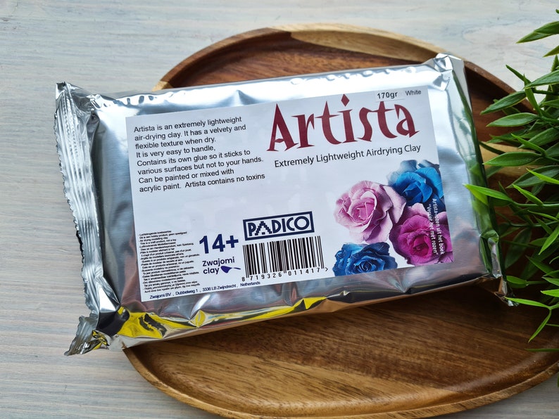May include: A silver package of white air-drying clay labeled "Artista Extremely Lightweight Airdrying Clay" with a barcode and the text "170gr White". The package features a floral design with pink, blue, and purple roses.
