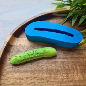 Könnte beinhalten: Eine blaue Silikonform in Form einer Gurke, neben einer grünen Plastikgurke auf einer Holzoberfläche.