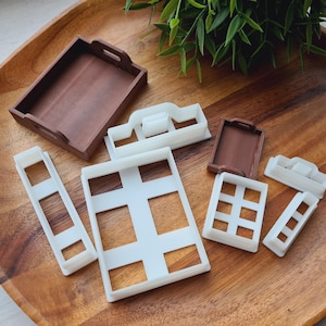 Può includere: Una collezione di formine per biscotti in plastica marrone e bianca di varie forme, tra cui vassoi rettangolari e disegni a forma di finestra. Le formine sono disposte su un vassoio di legno, con un fogliame verde sullo sfondo.