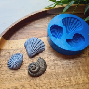 May include: A round blue silicone mold featuring three distinct seashell cavities, accompanied by three finished seashell castings in shades of metallic blue and dark bronze, displaying intricate ribbed and spiral textures on their surfaces.