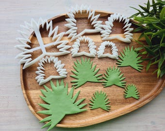 May include: A set of white plastic cookie cutters shaped like palm leaves and flowers on a wooden surface. There are also green fondant palm leaves of various sizes.