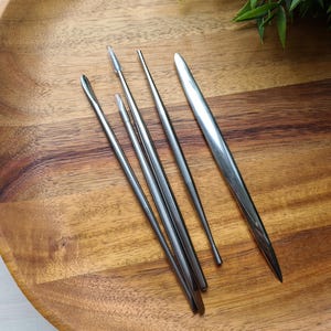 Könnte beinhalten: Ein Set aus vier silberfarbenen Metall-Modellierwerkzeugen mit glatten, spitz zulaufenden Enden. Die Werkzeuge unterscheiden sich in Dicke und Spitzenform, darunter spitze, abgerundete und flache Ausführungen, alle mit polierter Oberfläche.