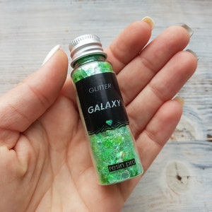 May include: A small glass bottle filled with green and iridescent glitter. The bottle has a black label with the words "GLITTER GALAXY" and "resin pro".