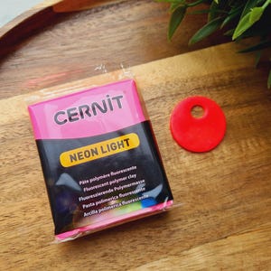 Peut inclure: Un paquet de pâte polymère fluorescente Cernit Neon Light rose et noir, et un petit disque rouge percé d'un trou. L'emballage contient du texte en plusieurs langues. Les articles sont sur une surface en bois.