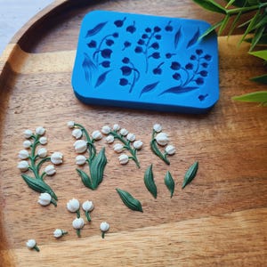以下が含まれることがあります： スズランのデザインの青いシリコンモールドと、いくつかの小さくて白と緑のスズランの装飾品。アイテムは木製のトレイに配置されています。