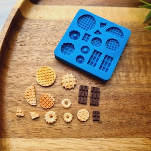 Może przedstawiać: Niebieska silikonowa forma do robienia mini wafli i batonów czekoladowych. Forma leży na drewnianej powierzchni, obok niej znajdują się mini wafle i batoniki czekoladowe.