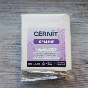 Może przedstawiać: Opakowanie białej gliny polimerowej, oznaczone jako "CERNIT OPALINE", z tekstem w kilku językach. Opakowanie zawiera 250g gliny i instrukcje pieczenia w temperaturze 110-130°C przez 10-30 minut.
