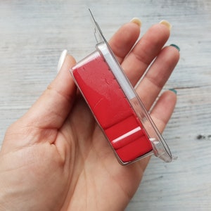 May include: A clear plastic package containing several rectangular, bright red objects. The package is held in a hand against a light-colored, wood-grain background. The objects appear to be a craft supply.