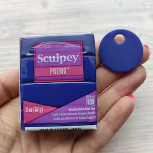 Peut inclure: Un paquet de 57 grammes d'argile à cuire au four Sculpey Premo de couleur bleu foncé. L'emballage est doté d'une étiquette rose avec le nom de la marque et des informations sur le produit. Un petit disque d'argile rond de couleur bleu foncé avec un trou au centre est visible en arrière-plan.