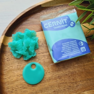 May include: A teal-colored polymer clay package labeled "CERNIT TRANSLUCENT" sits on a wooden tray, alongside a teal clay pendant and a teal clay flower. Green foliage is in the background.