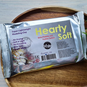 Puede incluir: Un paquete plateado de arcilla de modelado Hearty Soft Super Lightweight de color blanco. El paquete tiene una etiqueta blanca con texto negro que dice "Super Lightweight Modeling Clay", "Hearty Soft", "Extra Pliability and Feather Texture", "White", "Zwajomi Clay" y un código de barras. El paquete está sobre una superficie de madera.