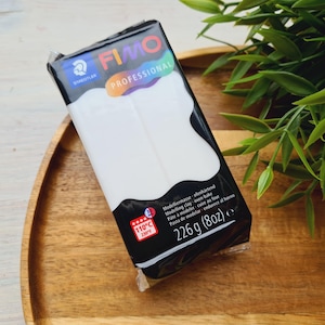May include: A package of white FIMO Professional modeling clay, 226g (8oz), with a black and white label. The label includes the text "FIMO PROFESSIONAL" and "STAEDTLER". The package is on a wooden tray with green foliage.