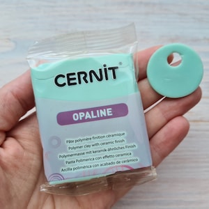 Może przedstawiać: Opakowanie gliny polimerowej Cernit Opaline z ceramicznym wykończeniem, w jasnym kolorze morskim, obok okrągłego kawałka w kolorze morskim z otworem. Opakowanie zawiera tekst w wielu językach.