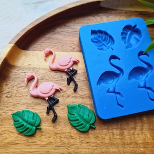 Könnte beinhalten: Eine blaue Silikonform mit zwei Flamingo- und zwei Blattdesigns. Die Form befindet sich auf einer Holzoberfläche mit zwei rosa Flamingo-Figuren und zwei grünen Blattfiguren.