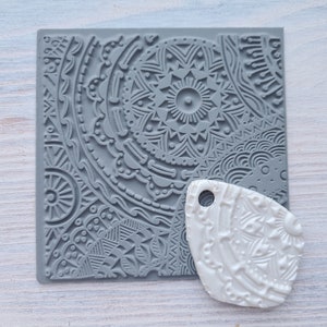 Può includere: Un timbro in silicone grigio con un design mandala dettagliato. Un ciondolo in argilla polimerica bianca con un design simile è accanto al timbro.