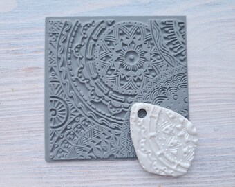 Pode incluir: Um carimbo de silicone cinza com um design de mandala detalhado. Um pingente de argila polimérica branca com um design semelhante está ao lado do carimbo.