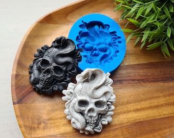 Könnte beinhalten: Eine schwarze, weiße und blaue Schädelform und -güsse. Das Schädeldesign zeigt eine Schlange, die sich um den Schädel windet. Die Form ist hellblau und die Gussteile sind schwarz und weiß. Die Objekte befinden sich auf einem Holztablett.