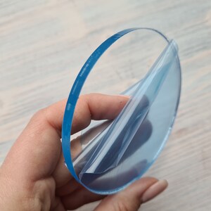 May include: A clear round piece of acrylic with a protective film being peeled off. The acrylic is translucent and has a slight blue tint.