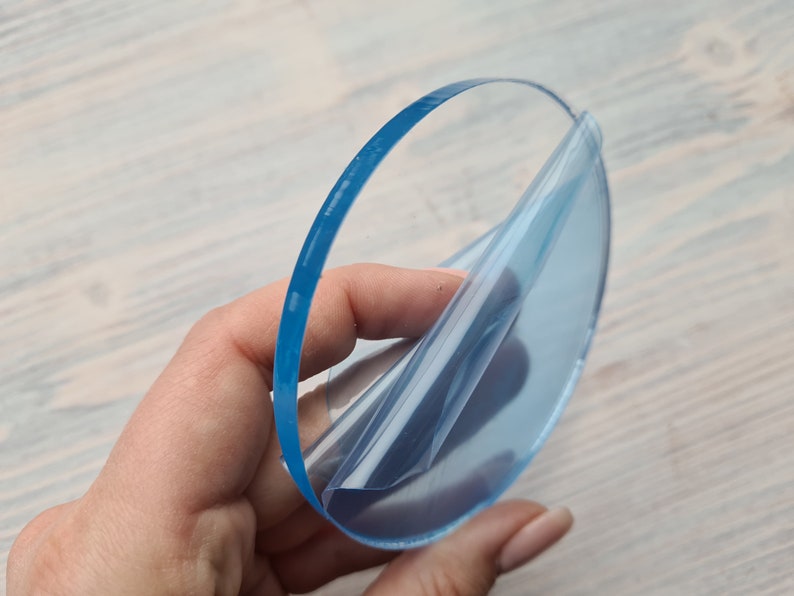 May include: A clear round piece of acrylic with a protective film being peeled off. The acrylic is translucent and has a slight blue tint.