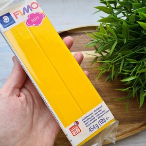 Puede incluir: Una mano sostiene un paquete de arcilla polim&eacute;rica FIMO SOFT amarilla. El paquete incluye el logotipo de Staedtler y la palabra "SOFT". Tambi&eacute;n indica que la arcilla es para hornear y pesa 454 g.