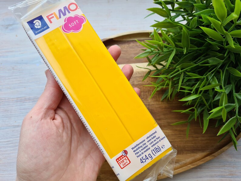 Puede incluir: Una mano sostiene un paquete de arcilla polim&eacute;rica FIMO SOFT amarilla. El paquete incluye el logotipo de Staedtler y la palabra "SOFT". Tambi&eacute;n indica que la arcilla es para hornear y pesa 454 g.