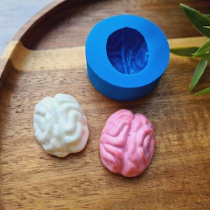 May include: A blue silicone mould shaped like a brain, with two finished brain-shaped sweets, one white and one pink, on a wooden surface.