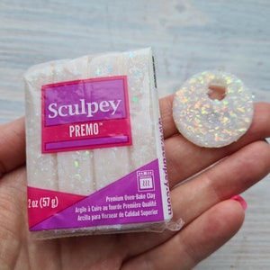 May include: A package of white Sculpey Premo oven-bake clay, 57 g, with a matching iridescent, donut-shaped clay piece. The package has a purple and pink label with the brand name and product details. The clay is premium quality.