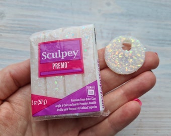 May include: A package of white Sculpey Premo oven-bake clay, 2 oz (57 g), with a matching iridescent, donut-shaped clay piece. The package has a purple and pink label with the brand name and product details. The clay is premium quality.