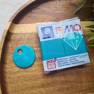 May include: A package of turquoise blue FIMO Effect modeling clay. The package has a white background with the brand name and product information printed in black. The clay is oven-bake and comes in a 57g package.