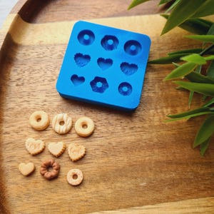 Könnte beinhalten: Blaue Silikonform mit 9 Vertiefungen zum Herstellen von Mini-Donuts, Herzen und anderen Formen. Die Form befindet sich auf einer Holzoberfläche mit mehreren Mini-Donuts und Herzen, die aus der Form hergestellt wurden.