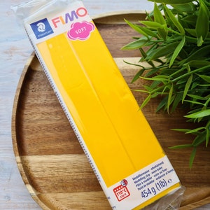 Puede incluir: Un paquete de arcilla de modelado amarilla FIMO SOFT, que pesa 454 gramos, se muestra en una bandeja de madera. El paquete tiene los logotipos STAEDTLER y FIMO SOFT, con texto que indica que es arcilla para hornear. La temperatura se muestra en grados Celsius.